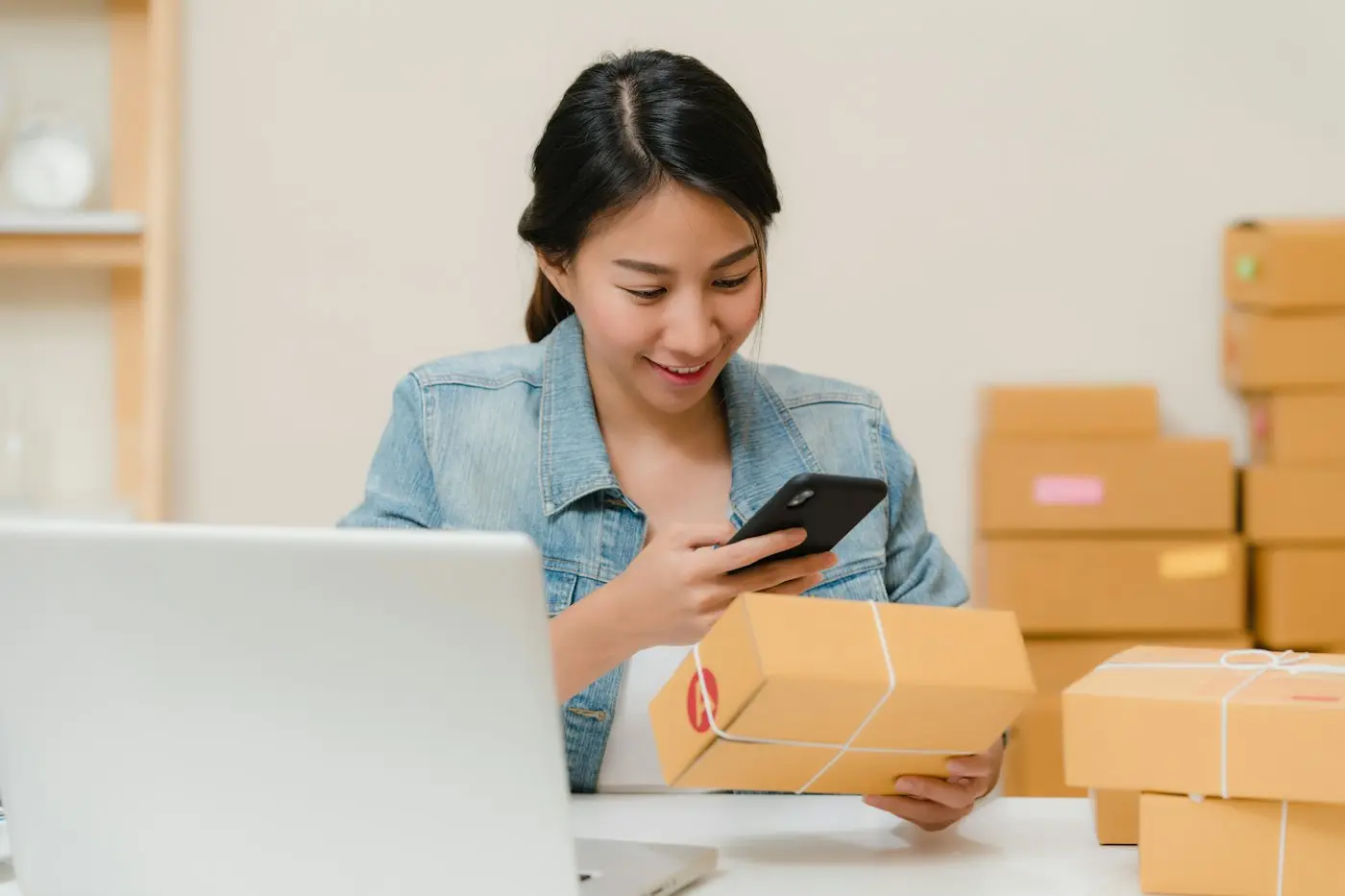 A woman checking a package on her phone next to her laptop and shipping boxes