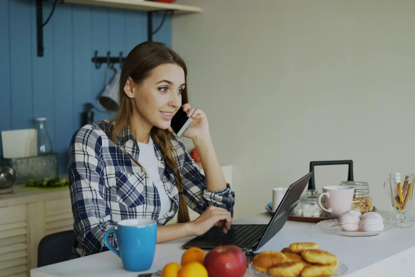 A smiling woman on a phone call at her kitchen table with her laptop open