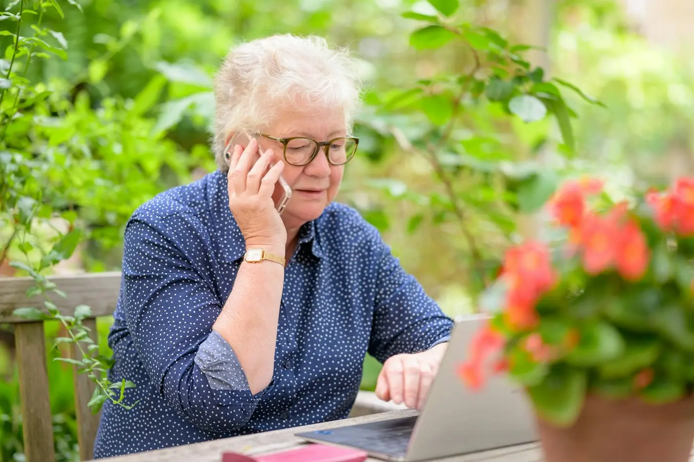 An older woman on a phone call while working on her laptop at a garden table